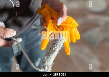 Artigiano con residui di vernice sulle mani e vestiti con un piccolo rullo di vernice e guanti in lattice ristrutturazione lavori sporchi formazione apprendistato p Foto Stock