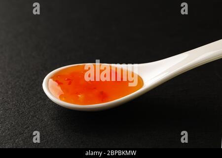 Dolce Chili sause in un cucchiaio di porcellana cinese bianco su una tavola di servizio in pietra nera. Salse e condimenti per carne, pesce e verdure. Vista frontale. Foto Stock