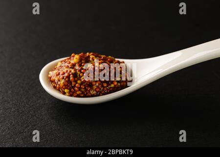 Senape a grani interi in un cucchiaio di porcellana cinese bianca su un asse di servizio in pietra nera. Salse e condimenti per carne, pesce e verdure. Vista frontale Foto Stock