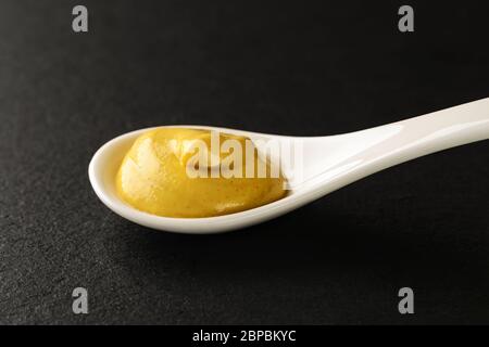Senape classica di digione in un cucchiaio bianco di porcellana cinese su un asse da servizio in pietra nera. Salse e condimenti per carne, pesce e verdure. Foto Stock