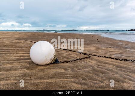 Vista ravvicinata di una boa bianca e della sua catena di ancoraggio, adagiata sulla sabbia sulla spiaggia, Bretagna, Francia, sotto un cielo nuvoloso Foto Stock