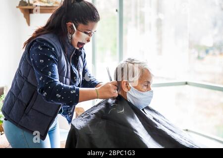 Donna anziana che ottiene un taglio di capelli a casa durante la pandemia Covid-19 indossare maschera facciale Foto Stock