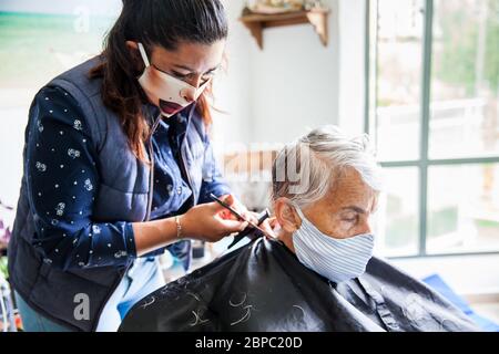 Donna anziana che ottiene un taglio di capelli a casa durante la pandemia Covid-19 indossare maschera facciale Foto Stock