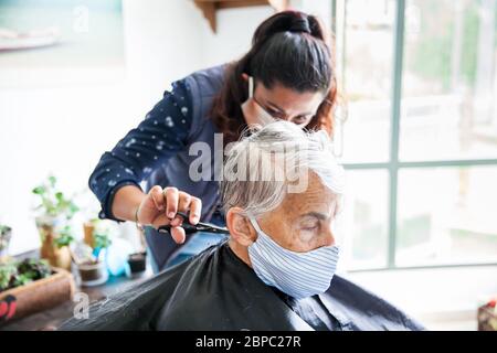 Donna anziana che ottiene un taglio di capelli a casa durante la pandemia Covid-19 indossare maschera facciale Foto Stock