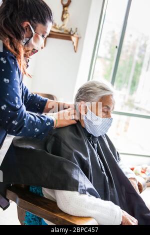 Donna anziana che ottiene un taglio di capelli a casa durante la pandemia Covid-19 indossare maschera facciale Foto Stock