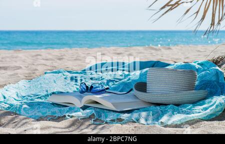 cappello, libro, asciugamano e occhiali da sole sulla spiaggia Foto Stock