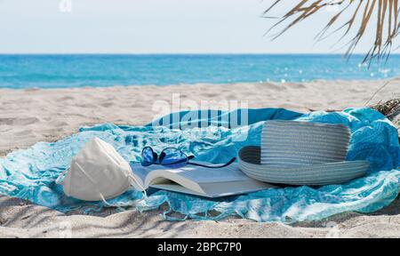 Maschera sanitaria, cappello, libro, asciugamano e occhiali da sole sulla spiaggia durante la pandemi19 Foto Stock