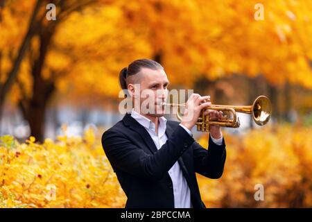 Giovane tromba europea bello in abiti formali che suona il suo strumento musicale tromba d'oro tra alberi con foglie gialle nel parco autunnale Foto Stock