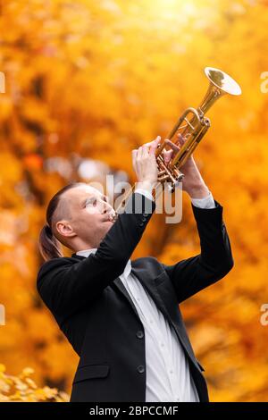 giovane bel muscolano e artista in costume nero e camicia bianca gioca tromba d'oro sullo sfondo delle foglie gialle d'autunno nel parco Foto Stock