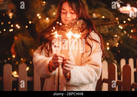 Felice ragazza adolescente che tiene scintillanti in piedi accanto al luminoso albero di natale in città Foto Stock