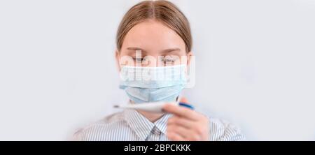 Primo piano ritratto, adolescente in maschera medica protettiva, controlla la temperatura termometro elettronico in mano. Banner orizzontale per foto per sito Web Foto Stock