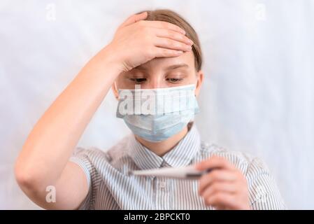 Primo piano ritratto, ragazza adolescente in maschera medica protettiva, tocca la fronte con la mano, tiene il controllo della temperatura sul termometro elettronico in Foto Stock