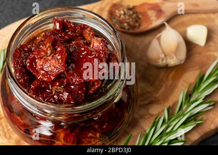 Pomodori secchi con aglio, rosmarino e spezie in un vaso di vetro su un tagliere di legno d'oliva. Foto Stock