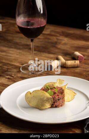 Tartare di bistecca con patatine su piatto bianco e tavolo in legno con bicchiere di vino e tappi Foto Stock