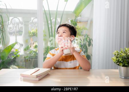 Un ragazzo asiatico beve latte mentre si siede alla scrivania dopo aver fatto i compiti. E-learning e concetto di istruzione. Foto Stock