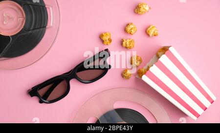 Composizione piatta con scatola di popcorn, occhiali 3d e bobine di pellicola su tavola rosa vista dall'alto. Guardare film online con il concetto di snack cinematografici. Foto Stock