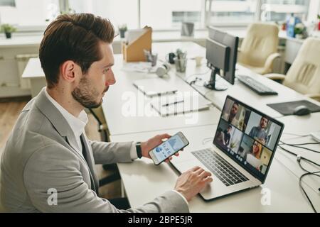 Giovane uomo d'affari con il portatore che lavora da solo in ufficio mentre collega i colleghi tramite videochat sul laptop durante l'epidemia di coronavirus Foto Stock