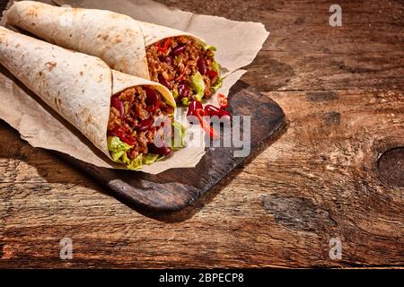Primo piano Still Life of Pair of Chili Stuffed Tex Mex Fajita avvolge in tortillas di farina grigliata e riposa su rustico tagliere su linguetta di legno Foto Stock