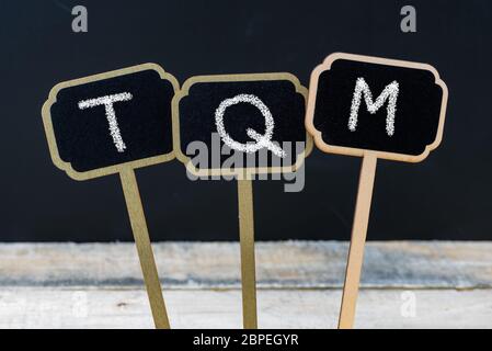 Acronimo commerciale TQM come Total Quality Management scritto con gesso su etichette in legno mini lavagna, lavagna defocata e tavolo in legno in backgroun Foto Stock