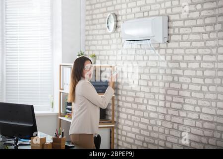 Giovane imprenditrice in piedi in ufficio utilizzando il telecomando del condizionatore aria Foto Stock
