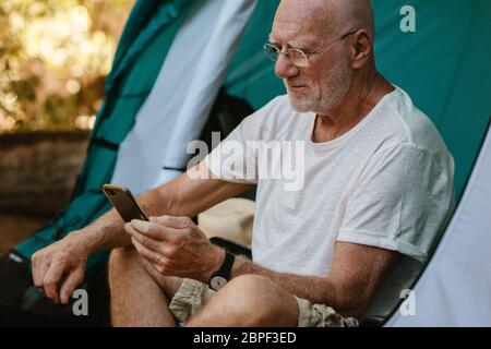 Uomo anziano seduto in tenda e utilizzando il suo telefono cellulare. Uomo maturo che campeggia in natura leggendo un messaggio di testo sul suo smartphone. Foto Stock