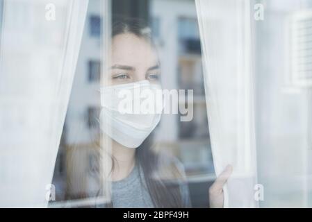 Giovane donna in maschera facciale guardando fuori la finestra. Rimanere a casa in quarantena. Foto Stock