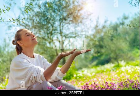 Bella donna felice seduta su prato di fiori nel giardino, godendo di sole di primavera luminoso, attiva vacanze di primavera in campagna Foto Stock