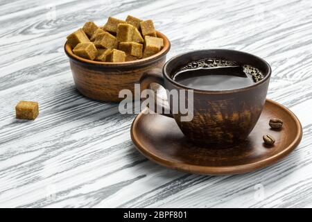 Caffè nero forte con schiuma in una tazza di ceramica e ciotola con cubetti di zucchero di canna su un tavolo di legno chiaro. Ci sono due grani di caffè su un piatto. Foto Stock