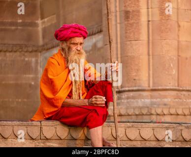 Varanasi, India - 13 novembre 2015. Un uomo caucasico, che indossa i colorati abiti tradizionali e la barba lunga di un sadhu indù, siede sui ghat. Foto Stock