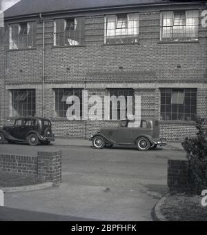 Anni '50, automobili storiche dell'epoca parcheggiata al di fuori di una fabbrica a due piani o edificio industriale, costruito in mattoni, con grandi finestre in metallo con piccoli pannelli, tipico del periodo, Inghilterra, Regno Unito. Foto Stock