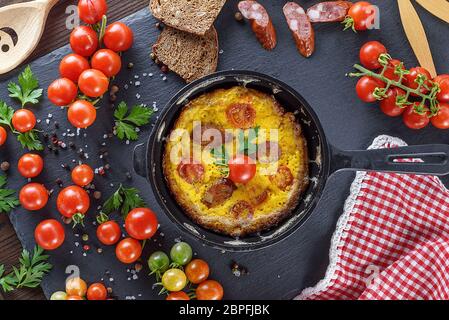 Fritti frittata di uova di pollo con rossi pomodori ciliegini e salsiccia affumicata in un tondo nero padella , vista superiore Foto Stock