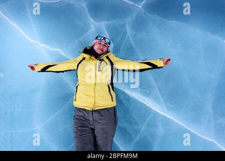 Ragazza felice in giacca nera gialla, pantaloni grigi, occhiali da sole blu a specchio e cappello rosa giace sul ghiaccio blu cracked del lago Baikal. Vista dall'alto Foto Stock