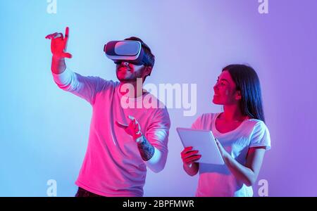 Ragazzo in realtà virtuale occhiali e mani in su, sorridente ragazza lo guarda e tiene tablet Foto Stock