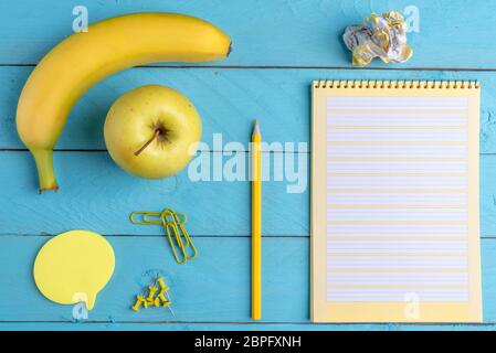 Il giallo delle forniture per ufficio e colazione su un legno blu scrivania in ufficio. Frutta fresca con oggetti di Office e di aprire il Blocco note Foto Stock