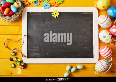 Layout di pasqua su giallo di tavoli in legno di uova colorate e caramelle intorno alla lavagna vuota tablet in cornice di legno per spazio di copia Foto Stock