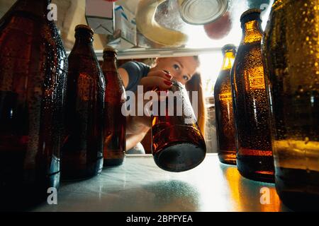 La donna caucasica prende la birra fresca rinfrescante dal frigorifero, vista interna dal frigorifero di mano che tiene la bottiglia. Alcohole, vita domestica, intrattenimento, concetto di bevanda. Cola fredda o bottiglie di birra. Foto Stock