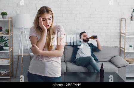 Donna infelice preme la mano per guancia, l'uomo guarda la TV e beve birra dalla bottiglia a casa Foto Stock