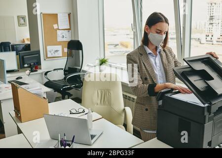 Giovane donna in ufficio in maschera di stoffa scansione documento in ufficio vuoto durante l'epidemia di coronavirus Foto Stock