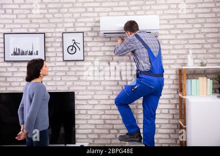 Giovane donna Guardando tecnico maschio riparare il condizionatore d'aria montati su un muro di mattoni Foto Stock