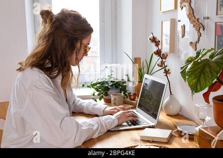 Giovane donna freelance/designer che lavora al computer da casa durante l'auto-isolamento a causa del coronavirus. Ambiente di lavoro accogliente circondato da piante Foto Stock