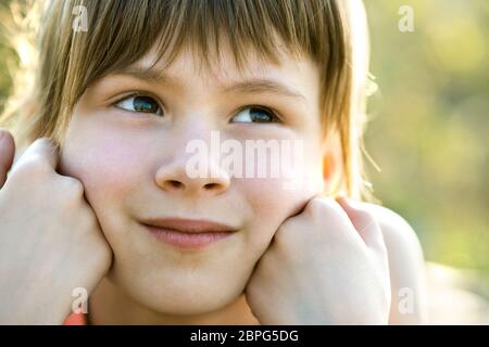 Ritratto di bella ragazza bambino con gli occhi grigi e lunghi capelli discreti appoggiandosi sulle sue mani sorridendo felicemente all'aperto su sfondo luminoso sfocato. Cute female Foto Stock