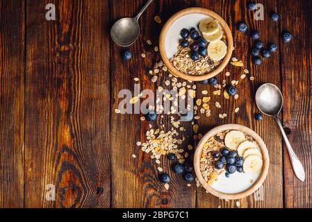 Due ciotola di muesli, Banana, mirtillo e yogurt greco. Ingredienti sparsi sul tavolo di legno. Vista da sopra e copia dello spazio. Foto Stock