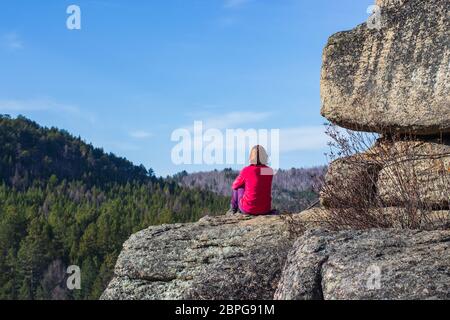 Una ragazza si siede in cima a una montagna da sola. Ammira la vista in una bella giornata di sole. Concetto di viaggio. Sfondo con spazio per il testo Foto Stock