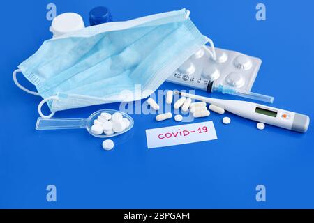 Maschera medica protettiva con pillole, termometro, farmaci, siringhe su scrivania blu. Virus pericoloso COVID-19. Concetto sanitario e medico. Foto Stock