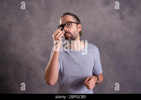 Giovane uomo barbuto ceaning i suoi occhiali di fronte a uno sfondo grigio Foto Stock