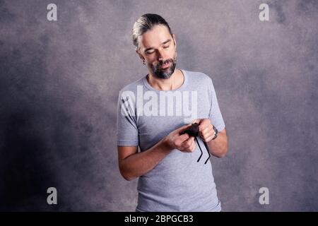 Giovane uomo barbuto ceaning i suoi occhiali di fronte a uno sfondo grigio Foto Stock