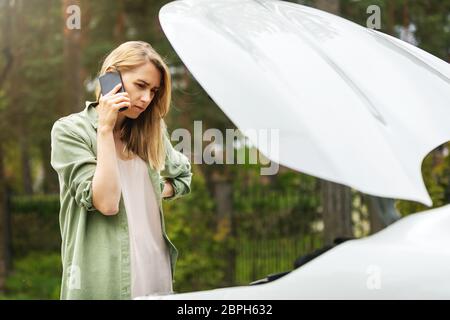 assistenza su strada - donna preoccupata in piedi davanti a auto rotta, guardando il motore e chiedendo aiuto Foto Stock