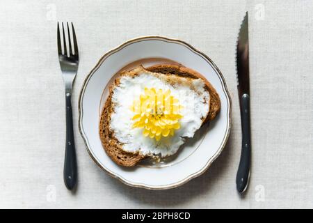 Immagine concettuale di fare colazione con toast fritti all'uovo. Fiore giallo di crisantemo invece di tuorlo servito su un piatto bianco Foto Stock