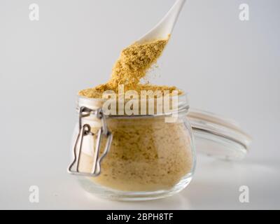 Lievito nutrizionale di fondo. Lievito nutrizionale inattivo che cade dal cucchiaio in vaso di vetro su sfondo bianco. Spazio di copia. Lievito nutrizionale è vegetale Foto Stock