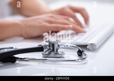 Close-up di un medico la mano utilizzando la tastiera del computer con uno stetoscopio sul bianco riflettente scrivania Foto Stock